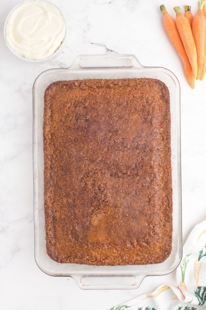 Baked carrot cake in a glass baking pan. The cake is unfrosted. Above the cake is a bowl of frosting and a bunch of fresh, peeled carrots.