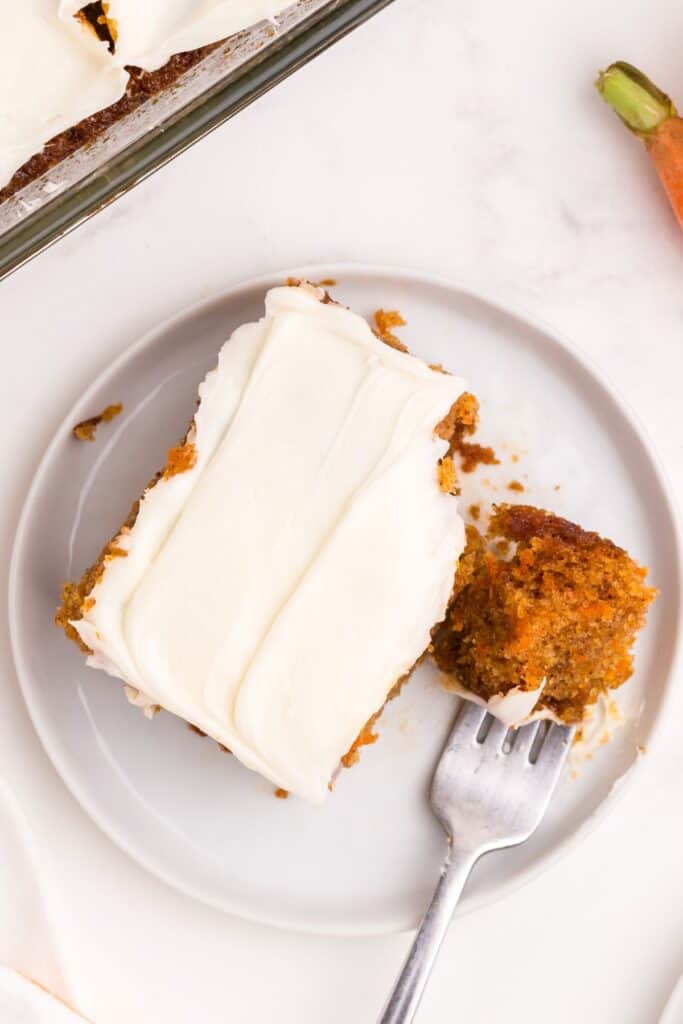 White plate with a serving of frosted carrot cake on it. A fork has cut away a bite of cake to show the interior.