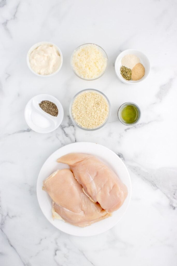 Ingredients for parmesan crusted chicken: chicken breasts, parmesan cheese, panko breadcrumbs, Italian seasoning, garlic powder, salt, black pepper, mayonnaise, olive oil, and lemon wedges.