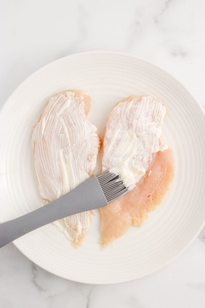 Prepped chicken breast coated in mayonnaise before dredging in parmesan cheese.