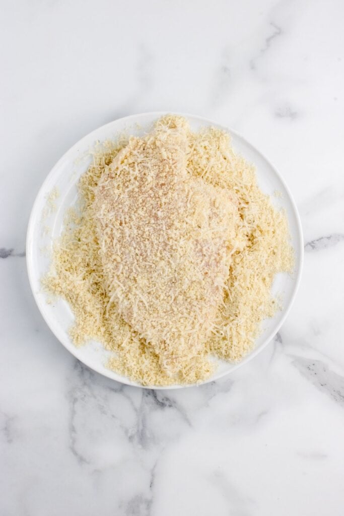 A white dinner plate with loaded with parmesan cheese for dredging chicken breast.