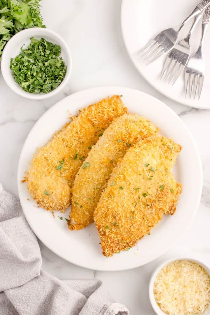 White serving plate with 3 pieces of parmesan chicken on it. Surrounded by dinner plates, forks, chopped parsley, and a small bowl of parmesan cheese.