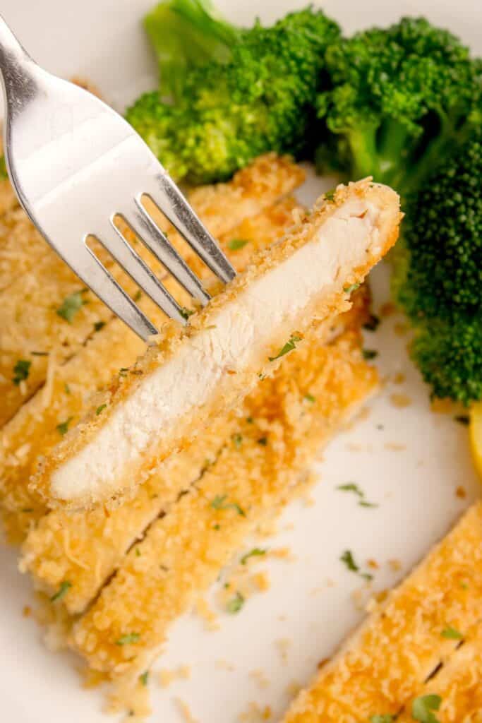 White dinner plate with sliced parmesan crusted chicken on it. A fork is lifting up a bite to show the interior of the chicken.