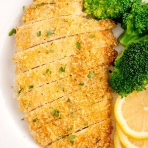White dinner plate with sliced parmesan crusted chicken, broccoli, and sliced lemon on it.