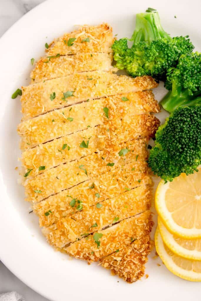 White dinner plate with sliced parmesan crusted chicken, broccoli, and sliced lemon on it.