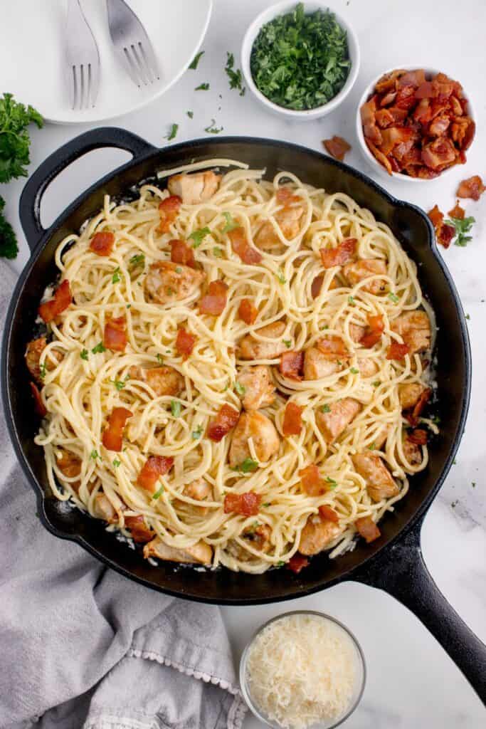 Cast iron pan of chicken carbonara. Pan is surrounded by small bowls of chopped bacon, parmesan, and parsley.