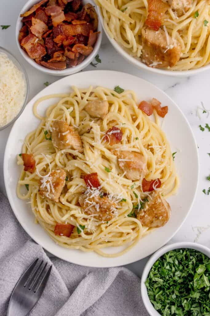 A white dinner plate with a serving of chicken carbonara on it. Plate is surrounded by a serving bowl of pasta, small bowls of chopped bacon, parmesan, and parsley.