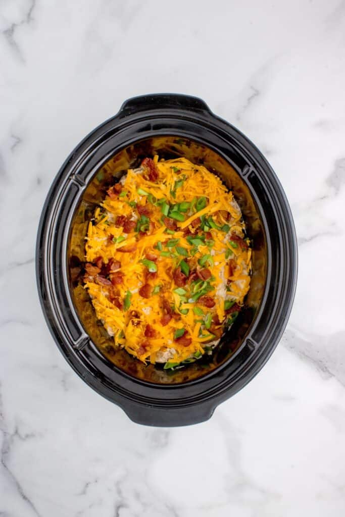 Slow cooker insert with cooked chicken. Chicken is shredded and layered with shredded cheddar cheese, bacon, and green onions.