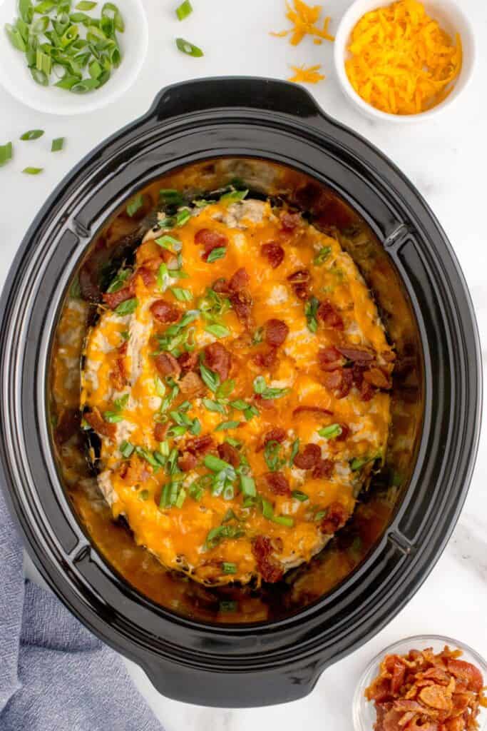 Crock pot insert with completed crack chicken. Chicken is shredded and layered with melted cheddar cheese, bacon, and green onions. The slow cooker insert is surrounded by small bowls of toppings: crumbled bacon, shredded cheddar cheese, and chopped green onions.