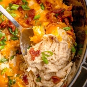 Crock pot insert with completed crack chicken. Chicken is shredded and layered with cheddar cheese, bacon, and green onions. A serving spoon is lifting a serving out of the insert.