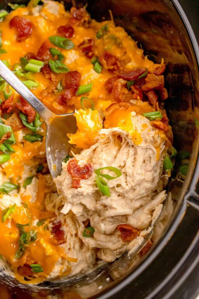 Crock pot insert with completed crack chicken. Chicken is shredded and layered with cheddar cheese, bacon, and green onions. A serving spoon is lifting a serving out of the insert.