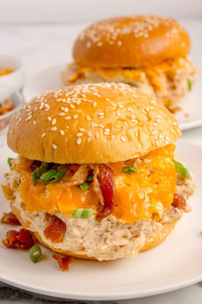 Sandwich with slow cooked chicken on a white dinner plate. Behind the plate is another white dinner plate with a chicken sandwich and two small bowls with shredded cheese and bacon.
