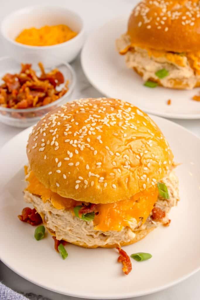 Sandwich with crack chicken on a white dinner plate. Behind the plate is another white dinner plate with a chicken sandwich and two small bowls with shredded cheese and bacon.