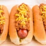 Close up view of a row of hot dogs on a serving platter with homemade chili and shredded cheddar cheese on top.