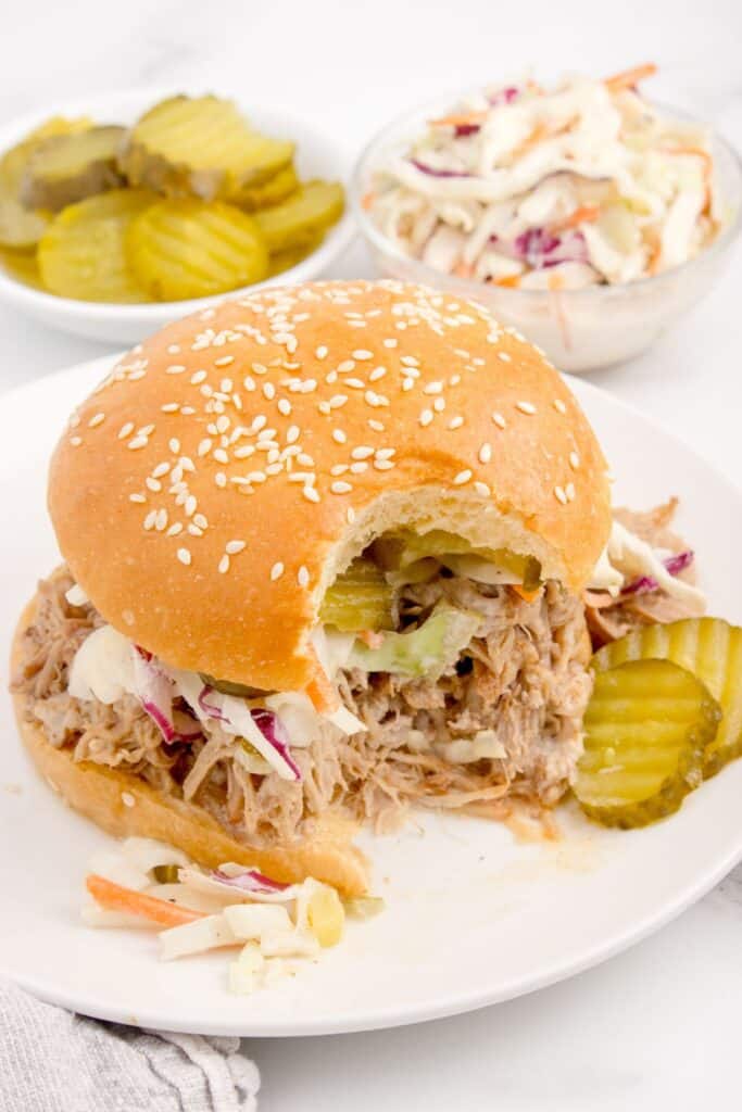 White dinner plate with pulled pork sandwich on it. A bite is taken out of the sandwich. Behind the plate is two small bowls with pickles and coleslaw.