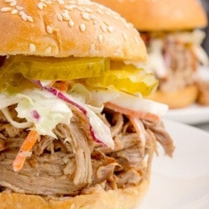 Pork sandwich layered with coleslaw and pickles on a white dinner plate.