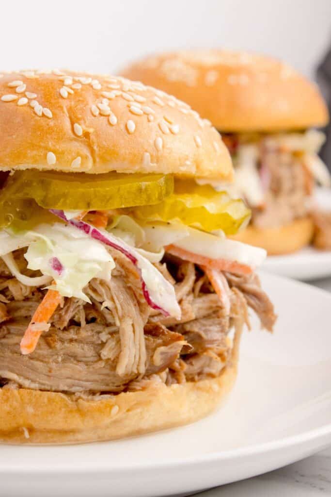 Pork sandwich layered with coleslaw and pickles on a white dinner plate.