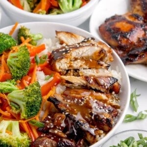 Teriyaki bowl surrounded by a bowl of veggies, a small glass bowl of teriyaki sauce, a plate of chicken thighs, and a small bowl of chopped green onions.