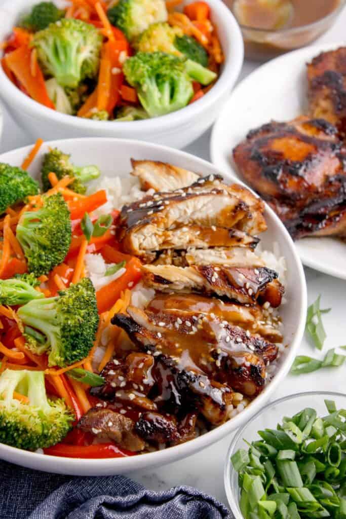 Teriyaki bowl surrounded by a bowl of veggies, a small glass bowl of teriyaki sauce, a plate of chicken thighs, and a small bowl of chopped green onions.