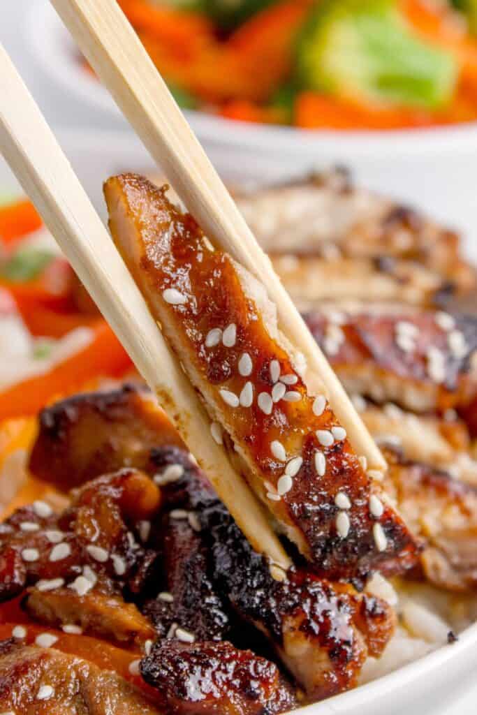 Up close view of teriyaki chicken, sprinkled with sesame seeds and being picked up by a set of chopsticks.