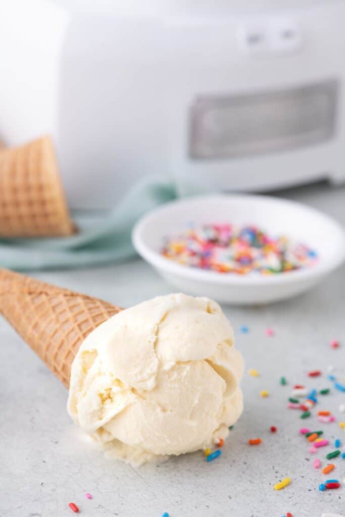 A sugar cone with a scoop of homemade vanilla ice cream on the table laying in a pile of sprinkles.