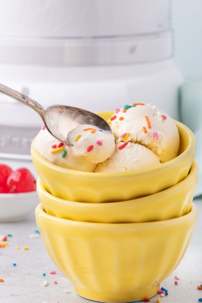 Bowl full of homemade vanilla ice cream with colorful sprinkles. A spoon is lifting a bite out of the bowl.