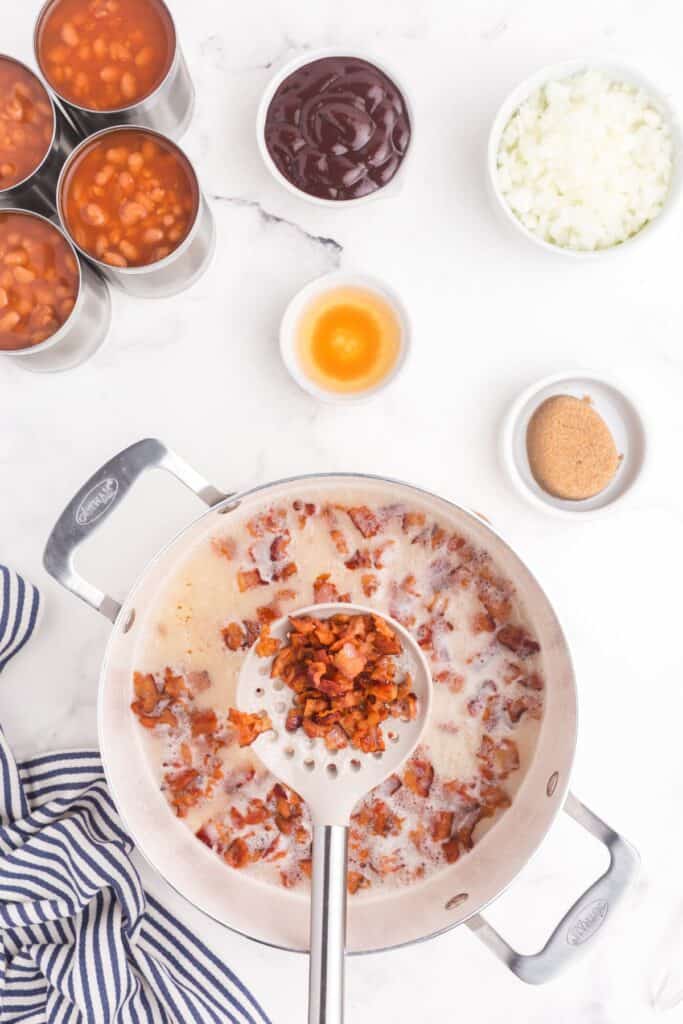 Pot with chopped bacon cooking in it next to 4 cans of baked beans, a small bowl of apple cider vinegar, a bowl of BBQ sauce, and a small bowl of packed dark brown sugar.