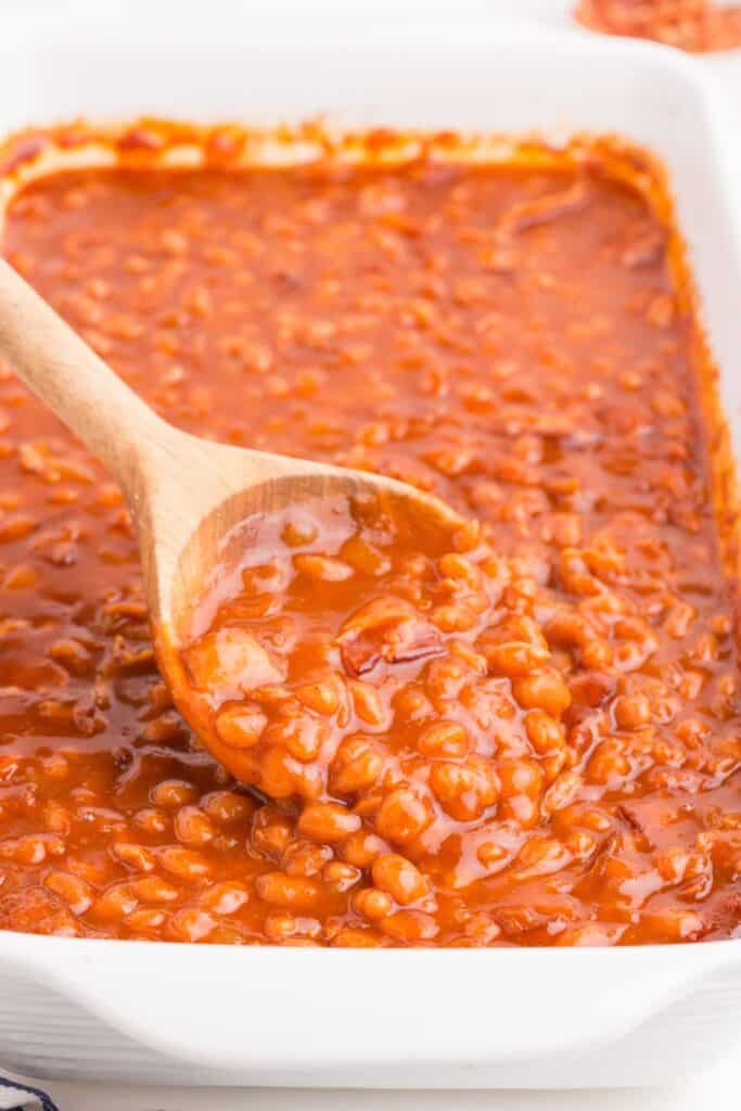 White baking dish with baked beans in it. A wooden serving spoon is lifting up a serving of beans.