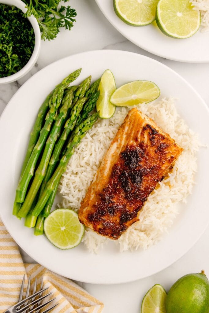 A white dinner plate with asparagus and white rice. A fillet of blackened mahi mahi is laid over top the rice. Plate is garnished with line wedges.