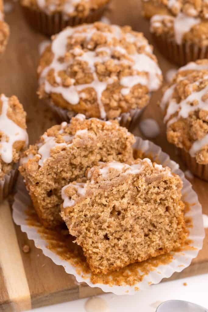 Rows of coffee cake muffins lined up on a wooden cutting board. The muffin in front have the paper wrapping peeled down and the muffin is sliced in half to show interior.