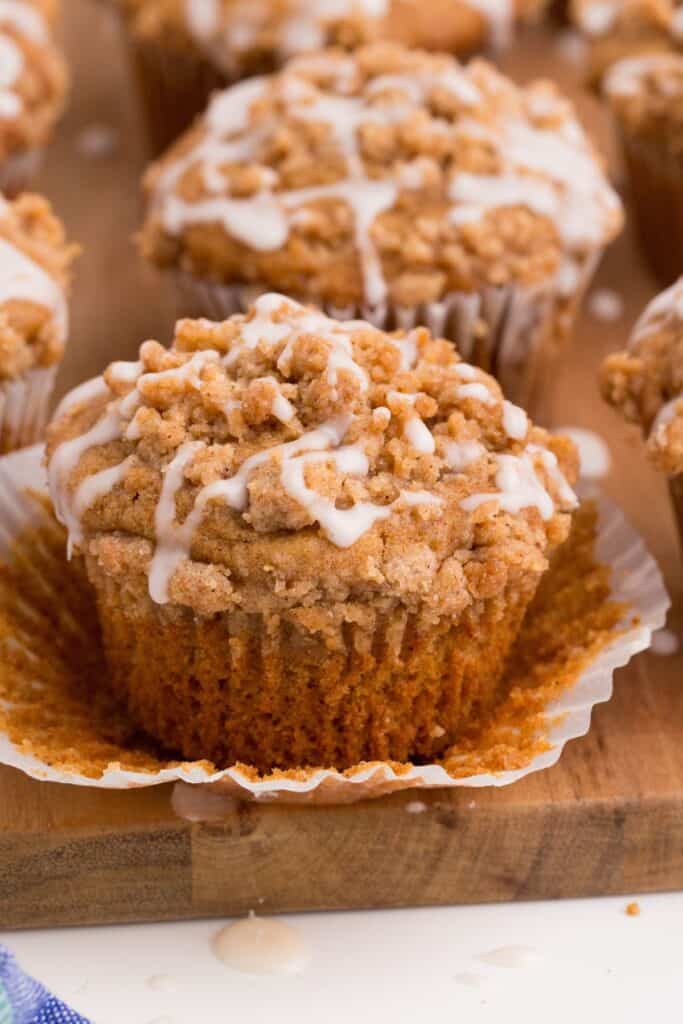 Rows of coffee cake muffins lined up on a wooden cutting board. The muffin in front have the paper wrapping peeled down.