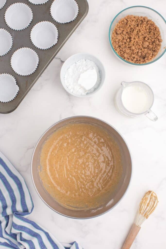 Mixing bowl with muffin batter in it. Behind the mixing bowl are ingredients for an icing, the streusel topping, and a paper lined muffin baking pan.
