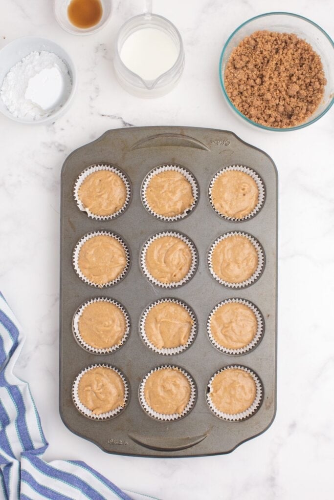 Unbaked coffee cake muffins prepped in a muffin baking pan. A medium sized bowl of streusel topping is above the pan, ingredients for icing are to the left of the bowl.