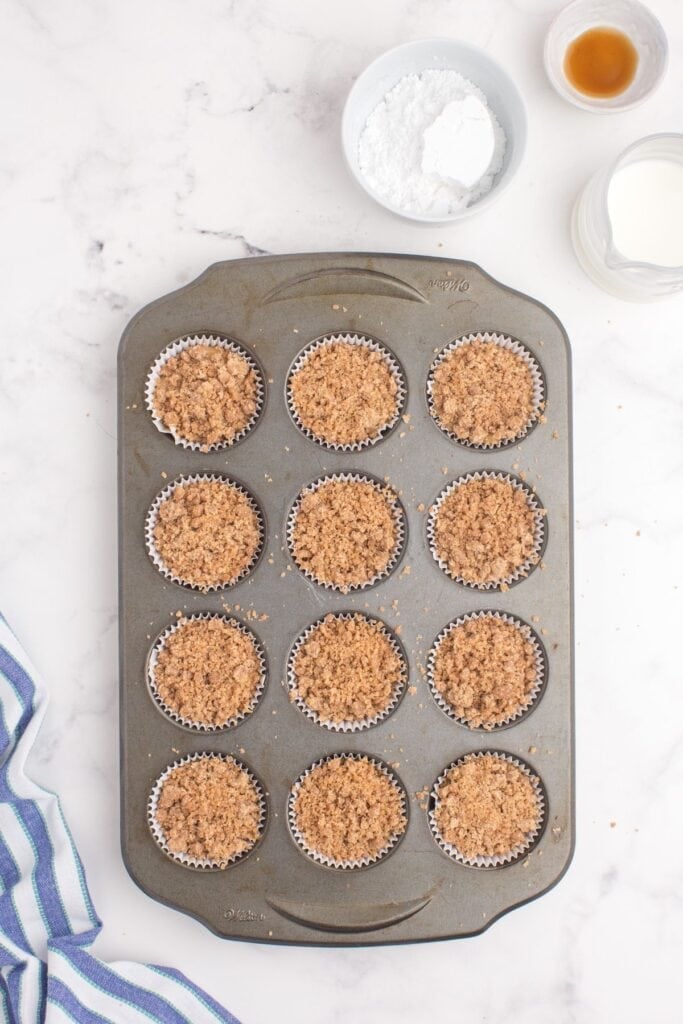 Unbaked coffee cake muffins with streusel are prepped in a muffin pan. Ingredients for icing (powdered sugar, vanilla, and milk) are arranged next to the pan.