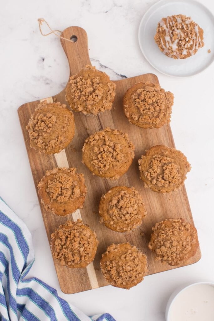10 Muffins lined up on a cutting board. They are not iced. There is one muffin on a plate next to the cutting board with an iced muffin.