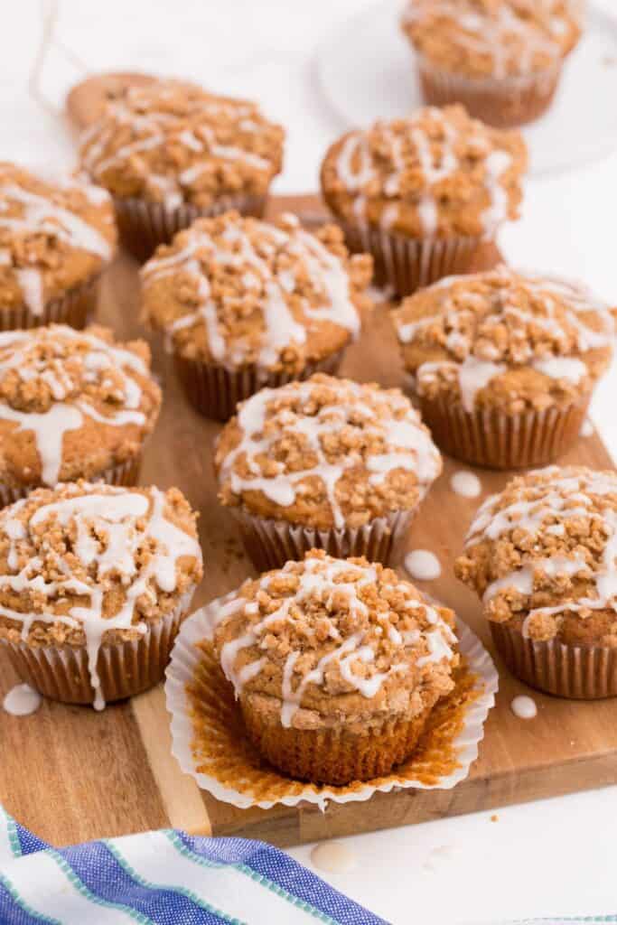 Rows of coffee cake muffins lined up on a wooden cutting board. The muffin in front have the paper wrapping peeled down.