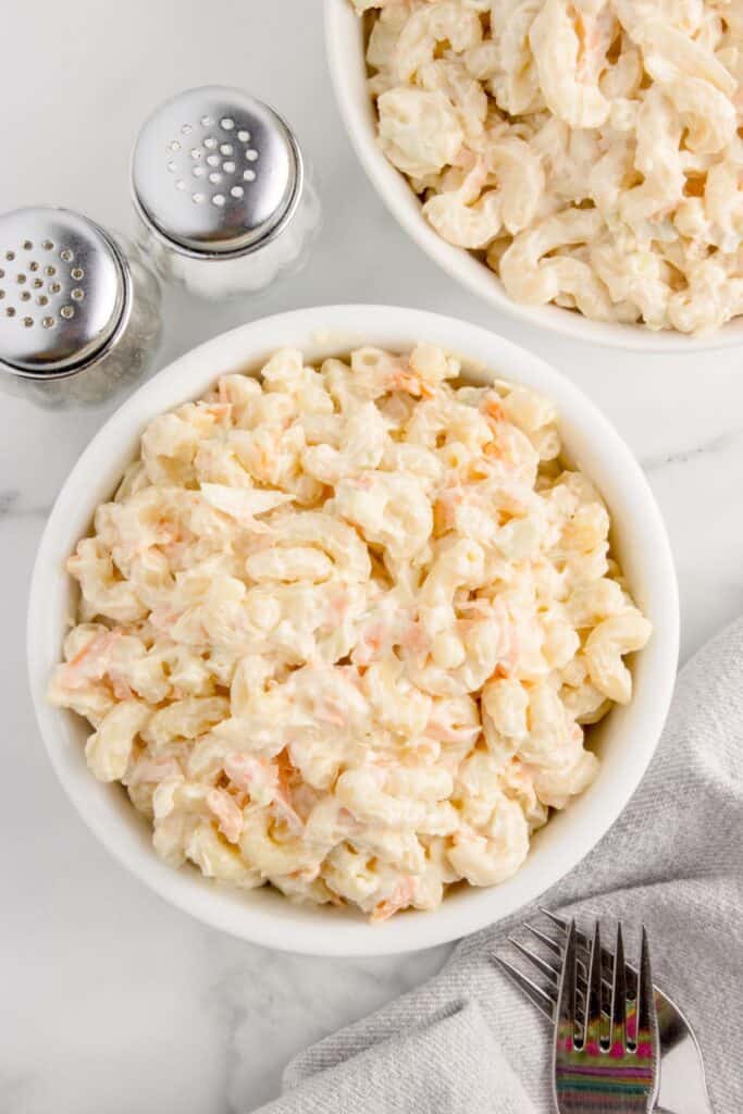 Two white bowls of macaroni salad. Bowls are flanked by a tea towel, forks, salt and pepper shakers.