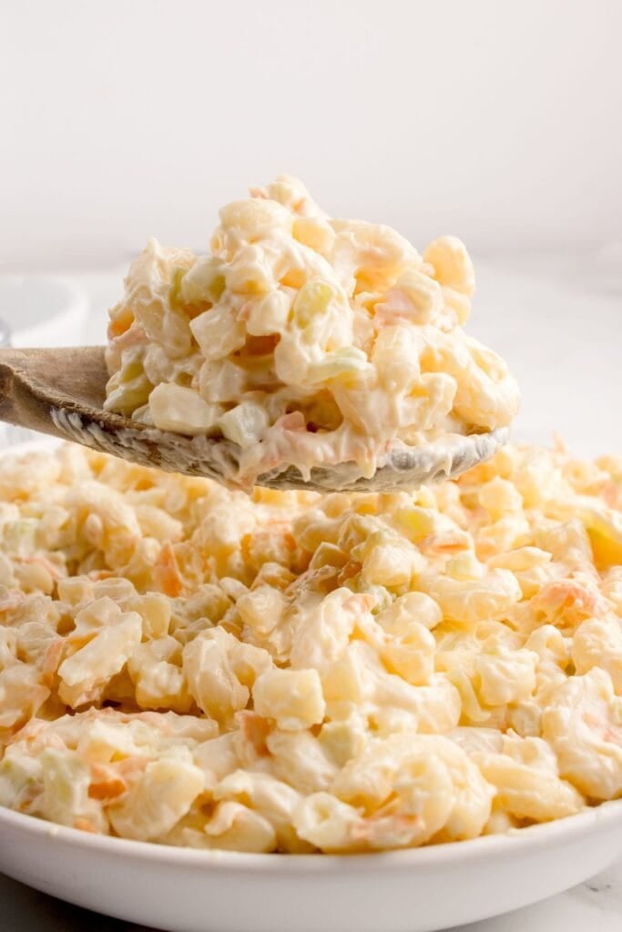 Creamy macaroni salad in a serving bowl with a wooden serving spoon lifting a portion above the bowl.
