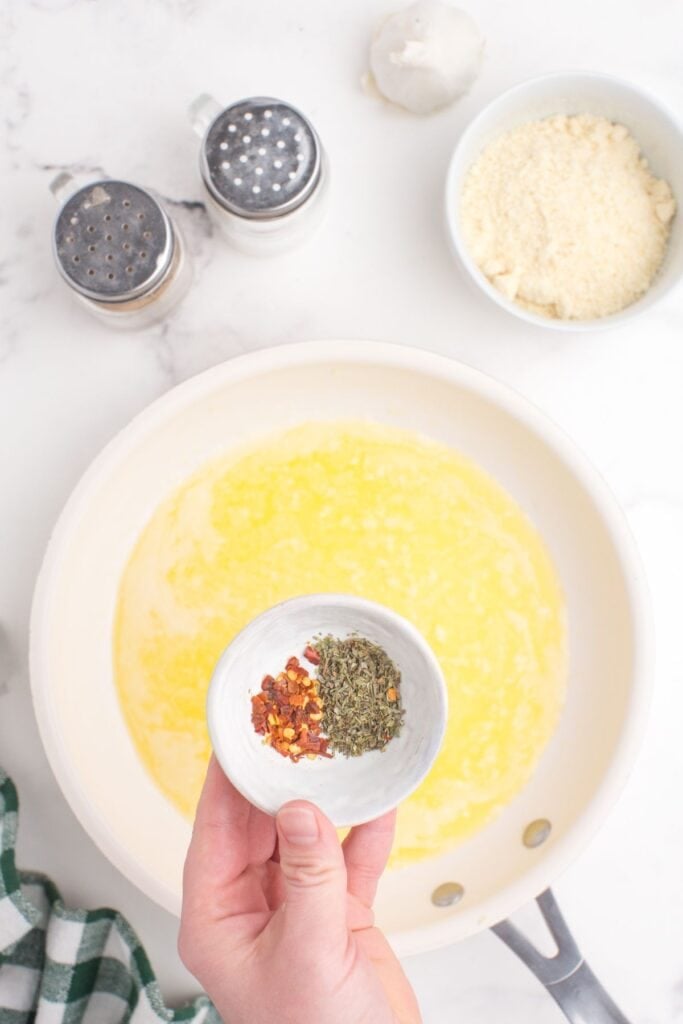 Melted butter and minced garlic in a skillet. Above the skillet is a small bowl of Italian seasoning and crushed red pepper. Surrounding the skillet is a bowl of grated parmesan cheese, a head of garlic, salt, and pepper.
