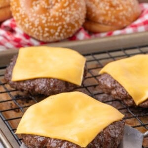 Cooked hamburger patties with melted cheese on a baking sheet. Hamburger buns and sliced vegetables are in the background.