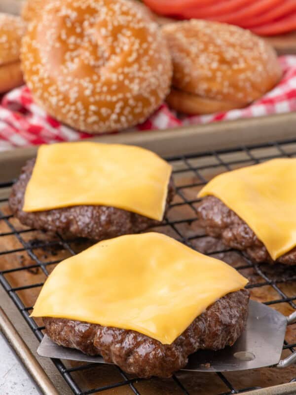 Cooked hamburger patties with melted cheese on a baking sheet. Hamburger buns and sliced vegetables are in the background.