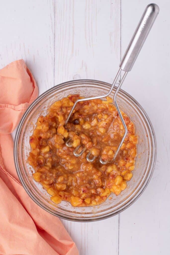 A glass bowl holds mashed and spiced peaches. A potato masher is in the bowl.