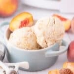 Ice cream scooped into a bowl garnished with a fresh peach slice. The bowl is surrounded by fresh whole peaches, peach slices, and an ice cream scoop.