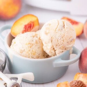 Ice cream scooped into a bowl garnished with a fresh peach slice. The bowl is surrounded by fresh whole peaches, peach slices, and an ice cream scoop.