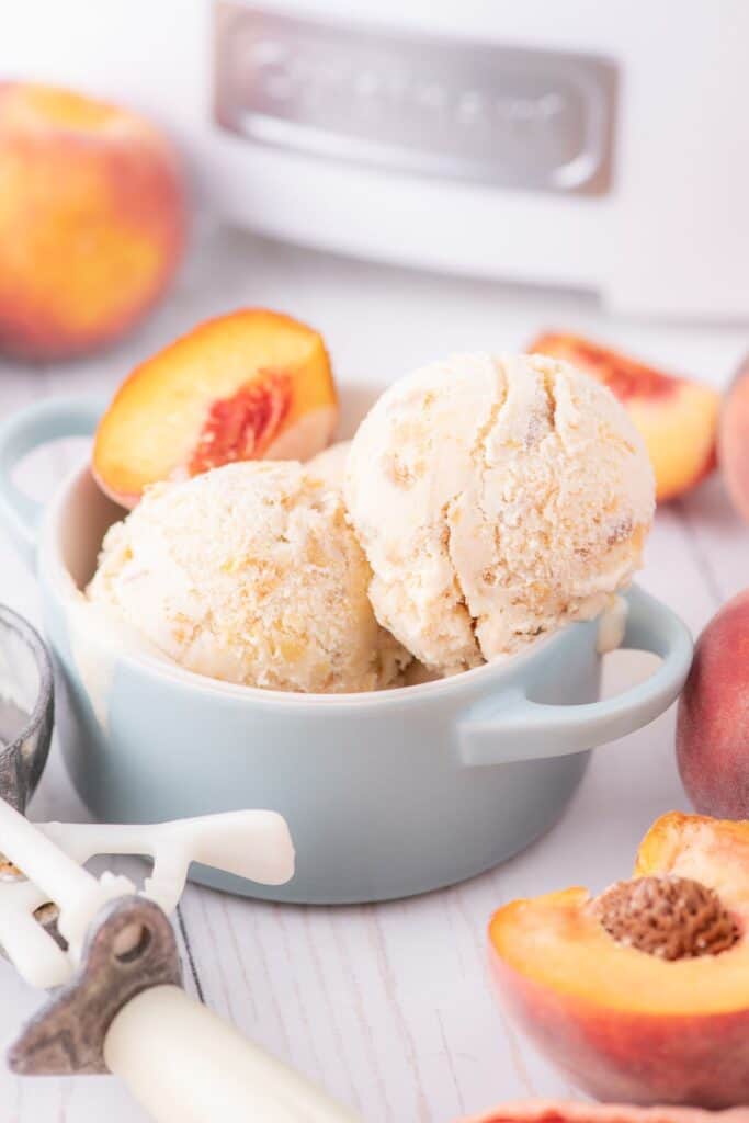 Ice cream scooped into a bowl garnished with a fresh peach slice. The bowl is surrounded by fresh whole peaches, peach slices, and an ice cream scoop.