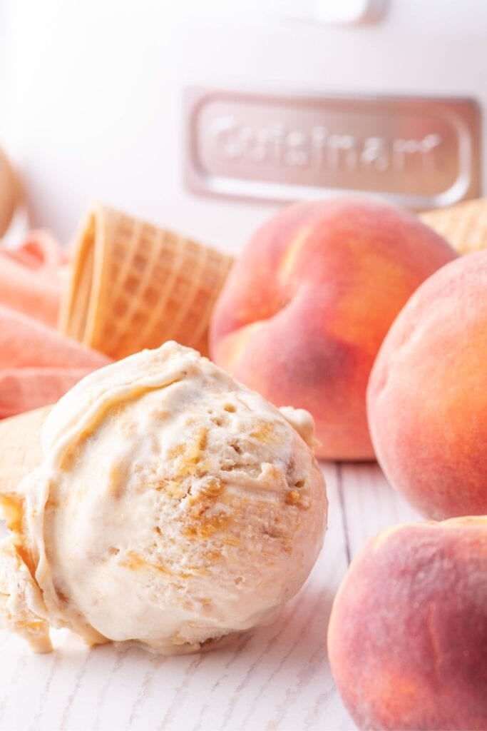 Peach ice cream on a sugar cone next to several fresh peaches.