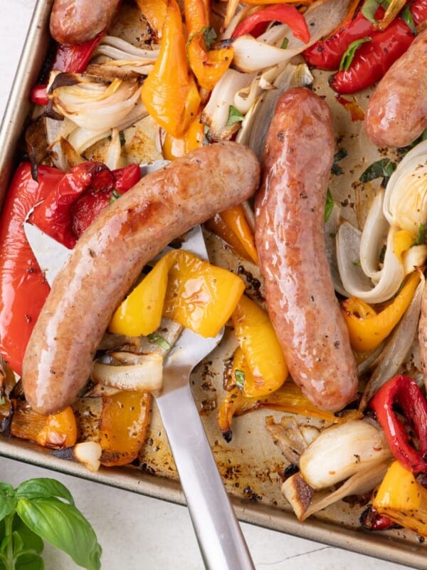 A serving spatula lifting up a serving of Italian sausage, bell peppers, and onions off a sheet pan.