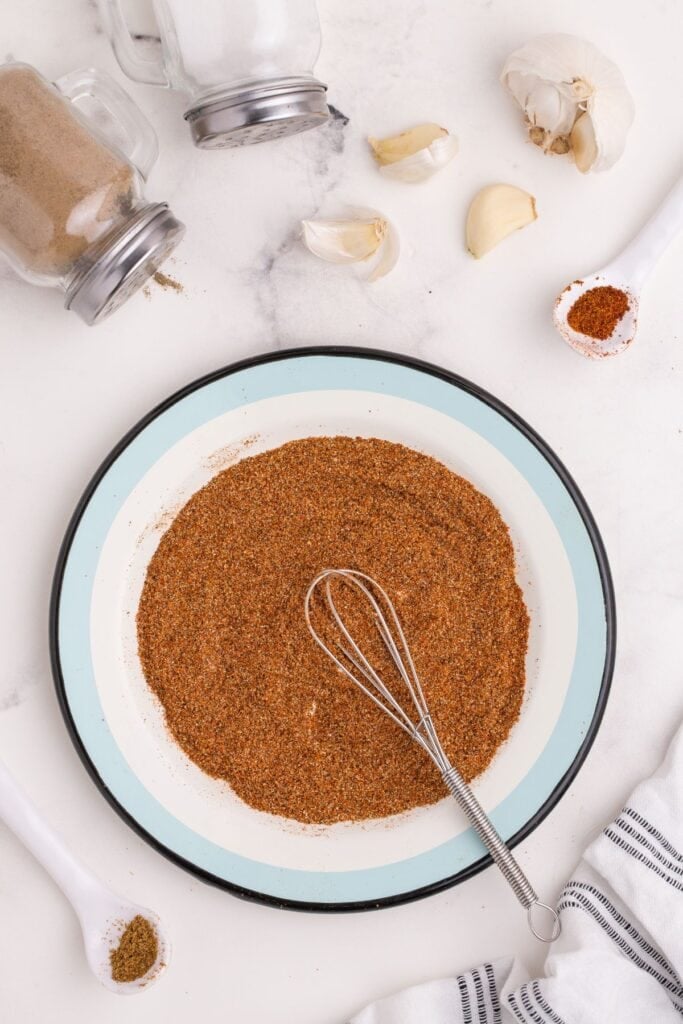 Chicken taco seasoning on a blue and white plate with a whisk. Plate is surrounded by a head of garlic, two white spoons, and a set of salt and pepper shakers.