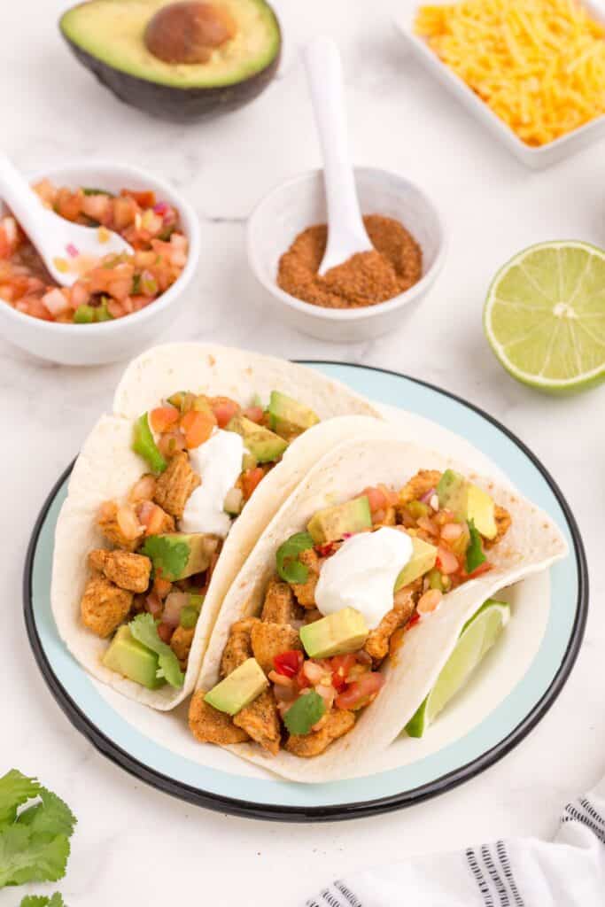 A blue and white plate with two chicken tacos in flour tortillas. Tacos are dressed with avocado, green onion, and sour cream. Behind the plate are small bowls of pico de gallo, homemade taco seasoning, shredded cheddar cheese, half a sliced lime, and half an avocado.