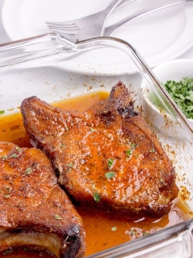 A glass baking dish with two baked pork chops with a sprinkle of parsley.